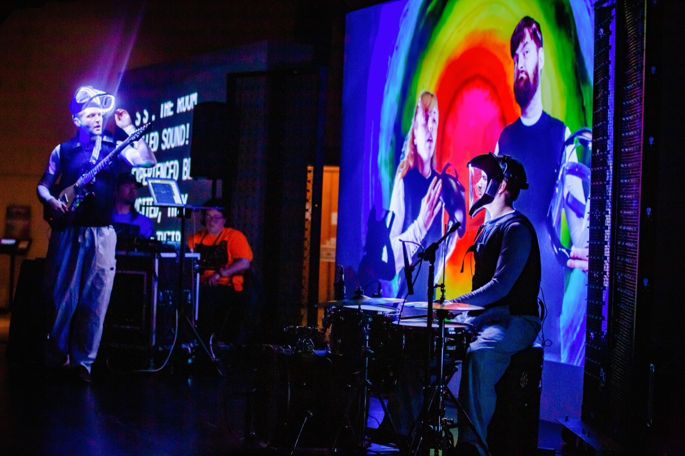 A band on a dark stage: a guitarist in a light-up helmet, a player behind a rack of electronics, and a drummer at a kit, with a screen behind them showing a rainbow field of concentric color rings and live video of two performers, and a caption screen at the left edge.