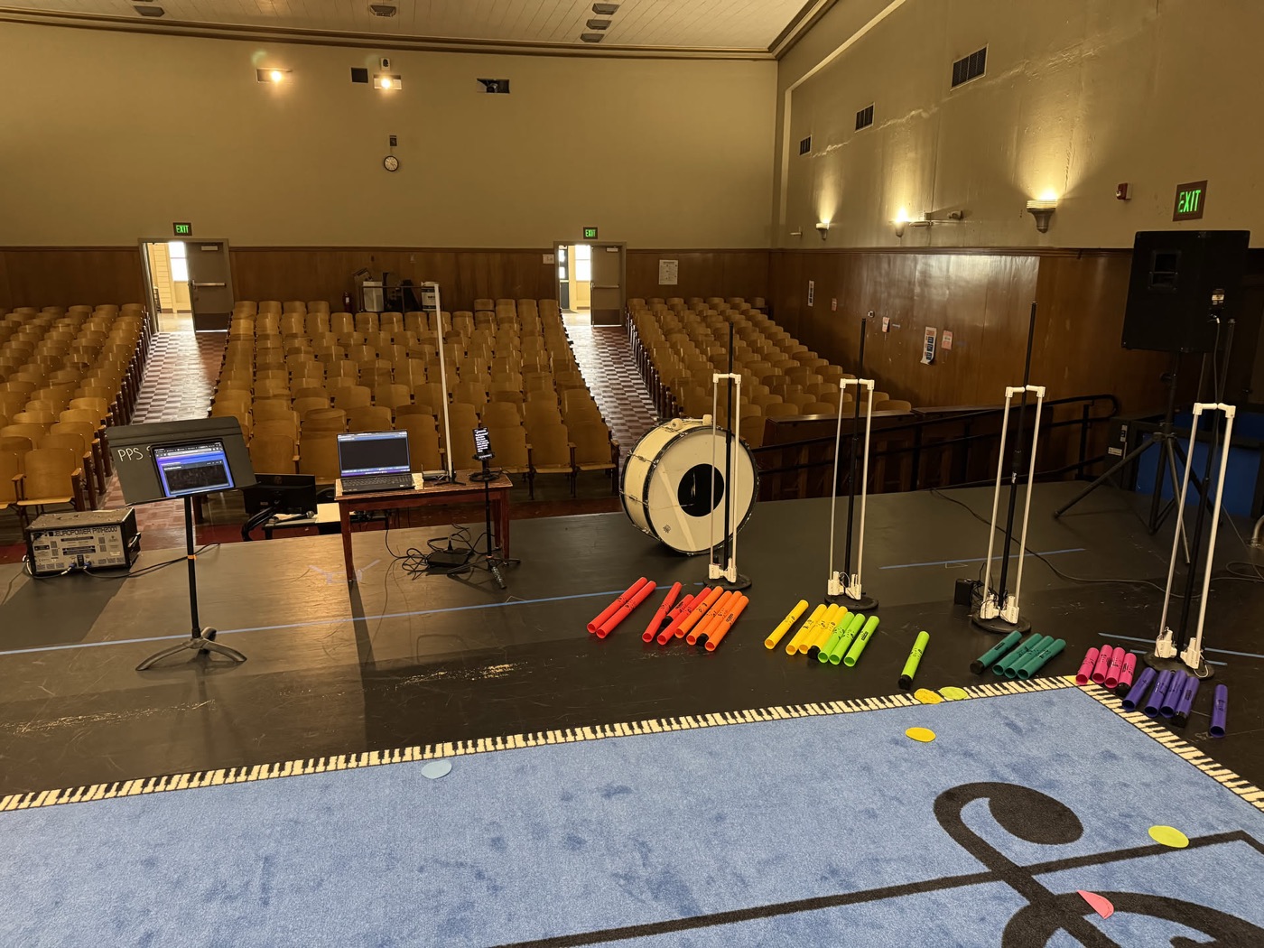 The workshop rig set up in an empty school auditorium: LED light tubes on stands, a bass drum, two laptops on a table, and color coded rows of tuned percussion tubes laid out on a floor mat printed with a treble clef and a piano keyboard border.
