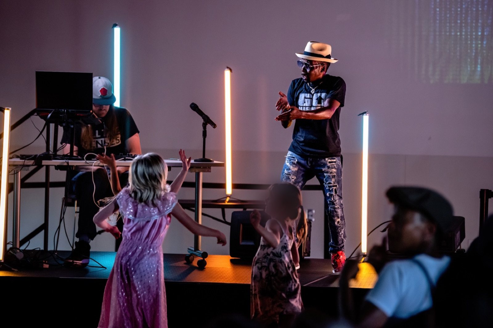 A performer in a straw hat on a low stage lit by vertical LED tubes, a laptop operator seated behind, and two children dancing in front of the stage with their backs to the camera.