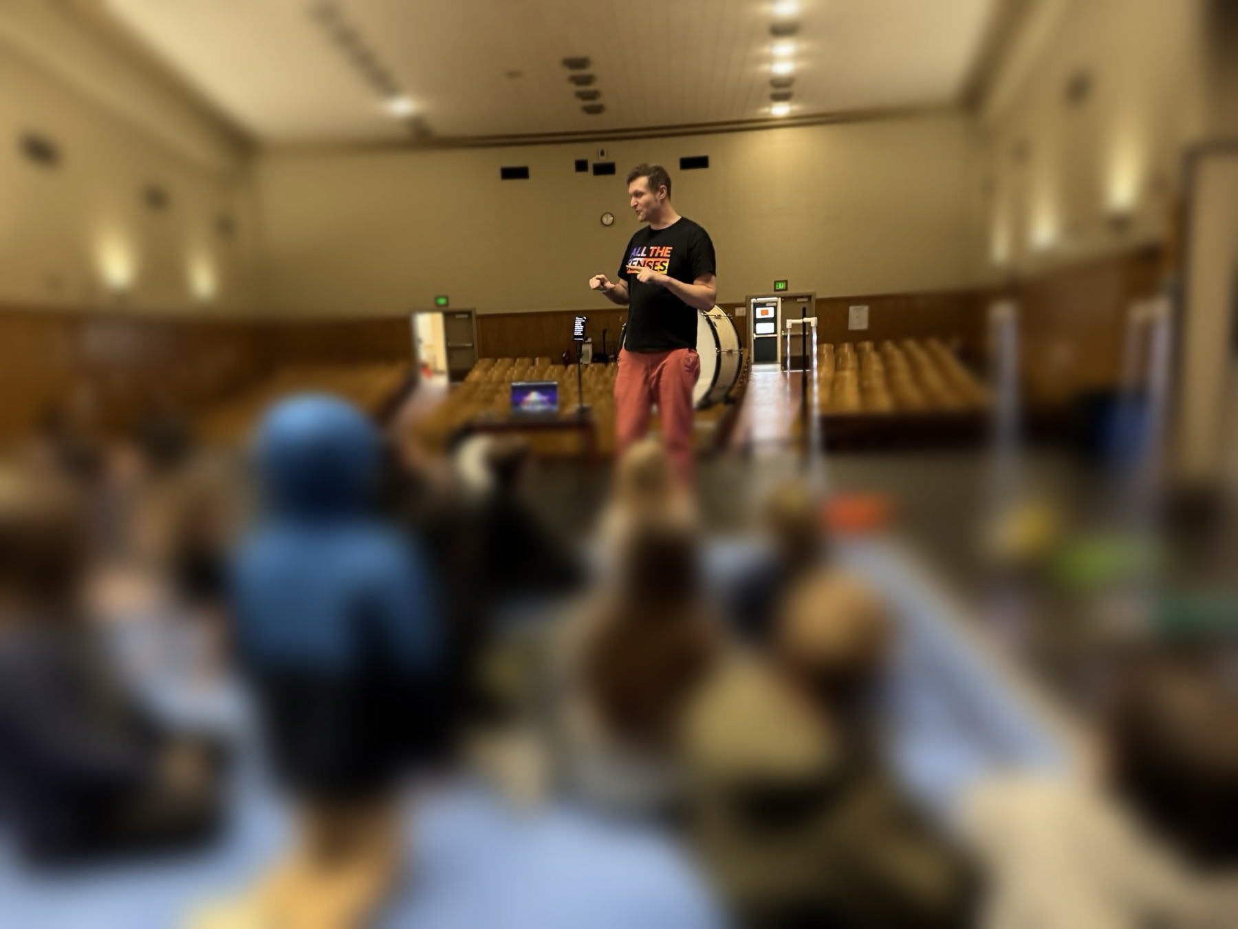 A workshop leader stands in a school auditorium beside a bass drum while children sit on the floor in the foreground, blurred and turned away from the camera.