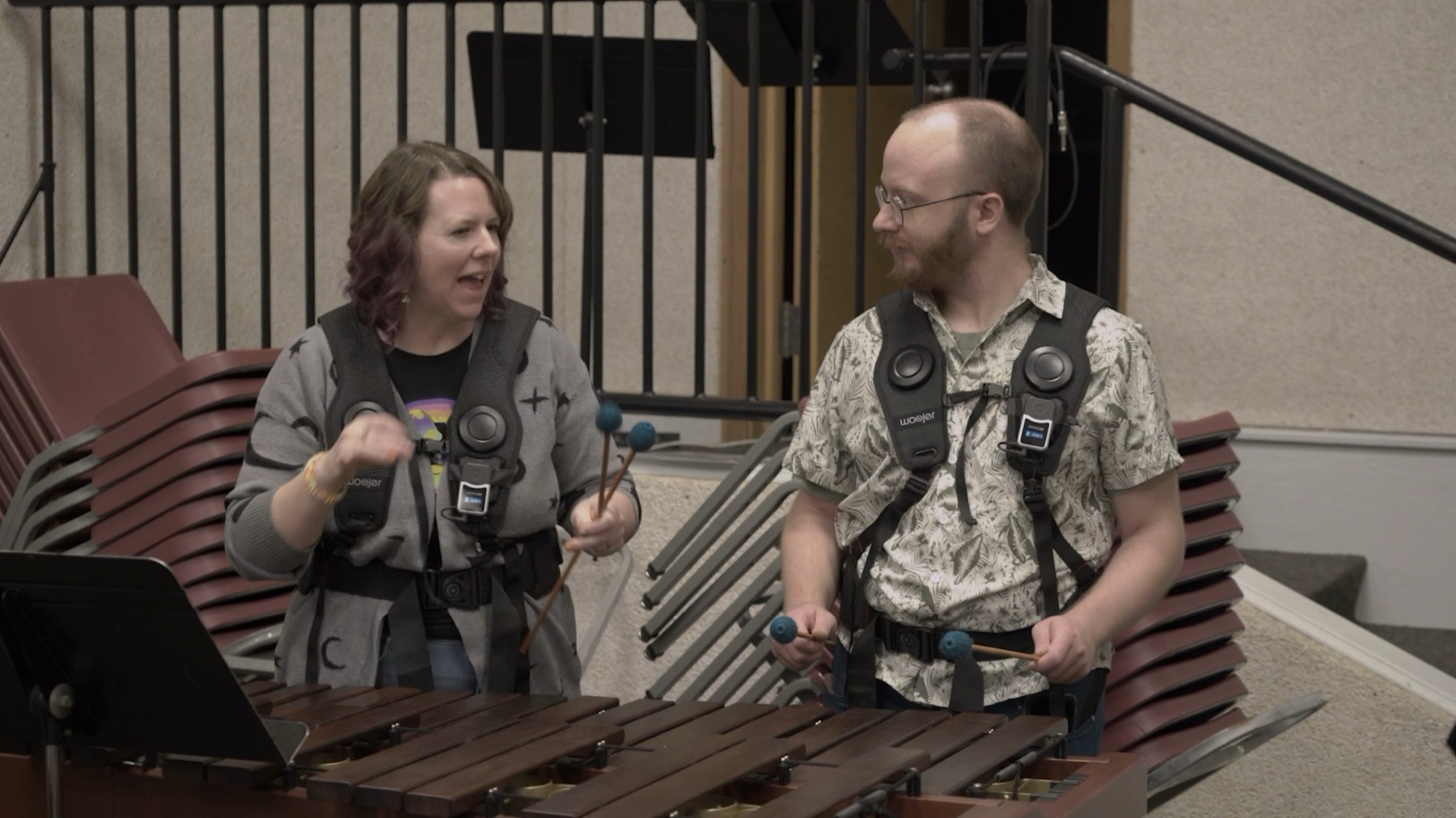 Two workshop participants wearing haptic vests stand at a marimba holding mallets, turned toward each other mid conversation.