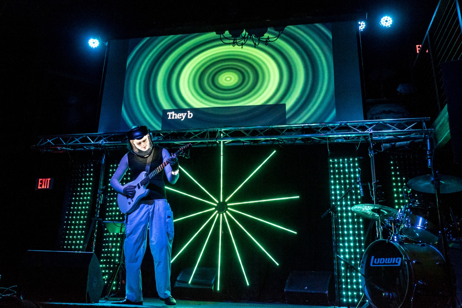 A performer in a helmet and haptic vest plays guitar on a dark stage while the sound is rendered behind as a large green starburst of concentric rings and radiating beams, flanked by walls of green LED panels, with a line of live captions above.