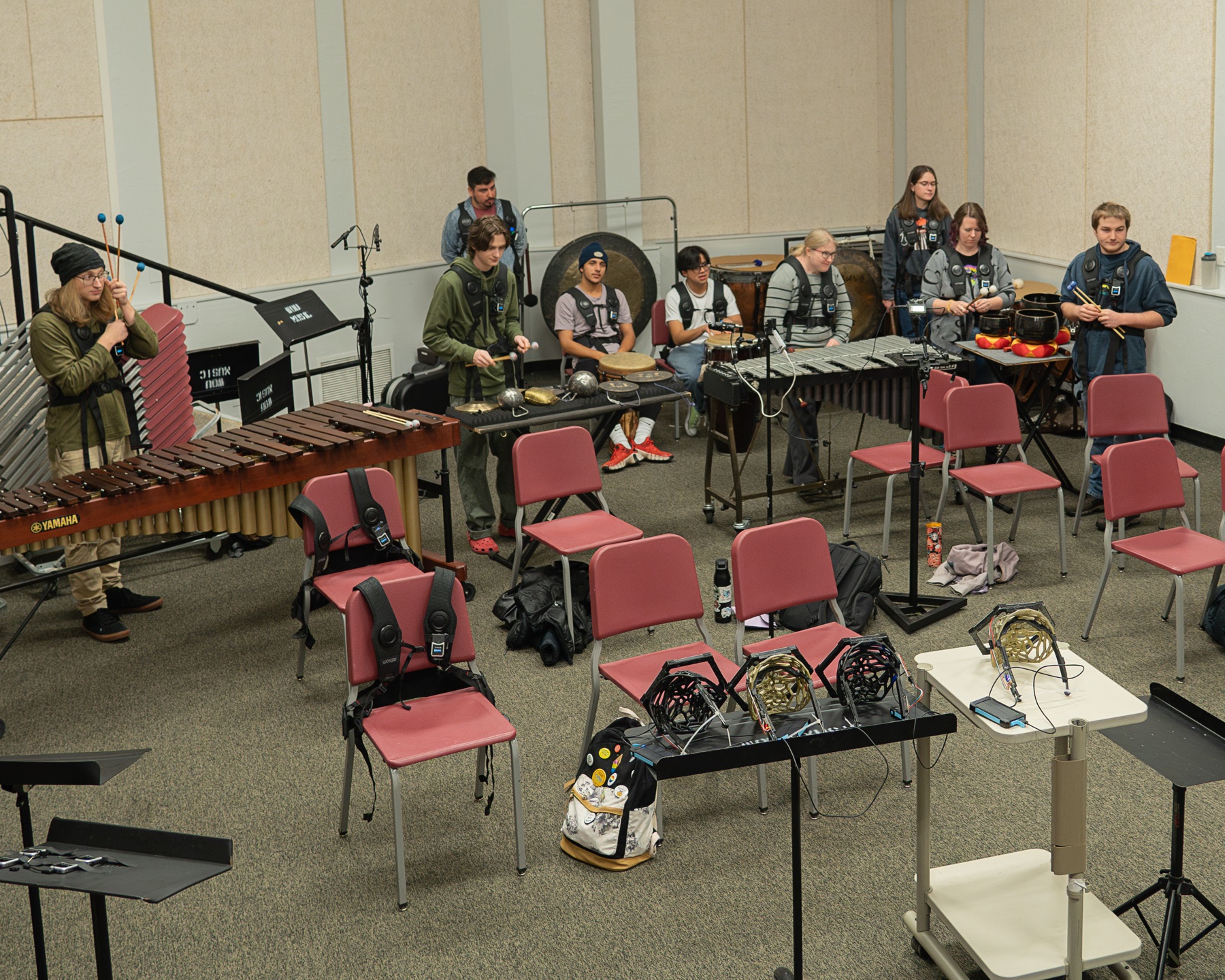 A university percussion ensemble spread around a rehearsal room with marimba, vibraphone, a large gong and hand percussion, most of the players wearing haptic vests over their clothes.