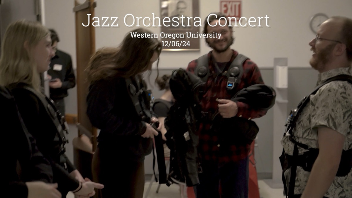 Four people stand talking in a venue lobby before a concert, each wearing or holding a haptic vest, with spare vests draped over one person's arm ready to hand out. Overlaid title text reads Jazz Orchestra Concert, Western Oregon University, 12/06/24.