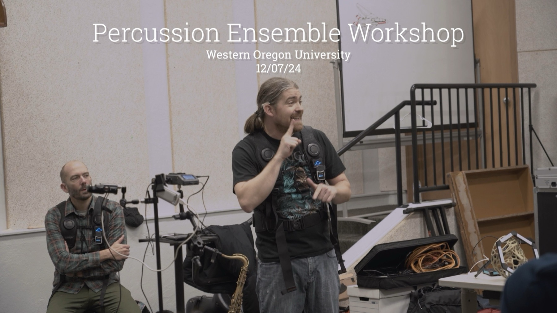A workshop leader wearing a haptic vest signs to a room while a seated colleague in another vest watches, surrounded by percussion gear, stands and cases. Overlaid title text reads Percussion Ensemble Workshop, Western Oregon University, 12/07/24.