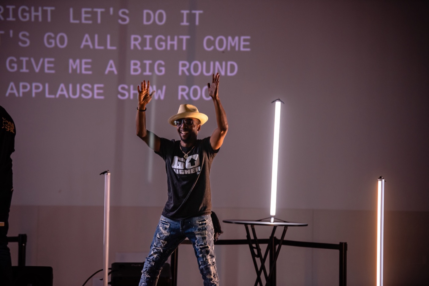 A performer in a straw hat and sunglasses grins with both arms raised on a low stage, in front of a wall filled with large projected capital-letter captions, with lit white and orange light tubes on stands beside him.
