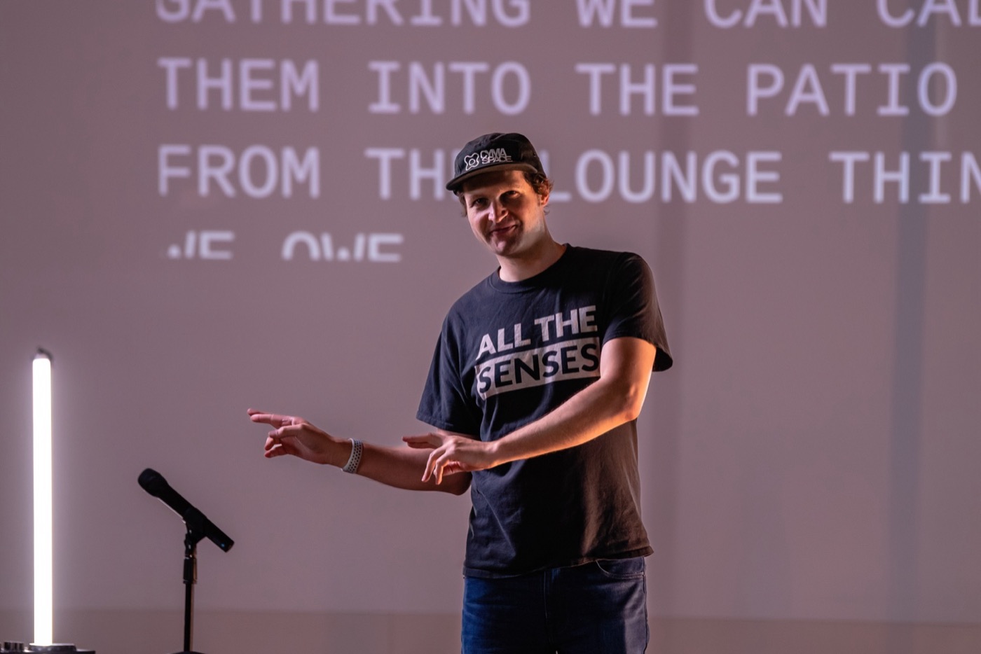 A person in a dark cap and a T-shirt reading All The Senses signs with both hands beside a microphone stand, with projected capital letters filling the wall behind.