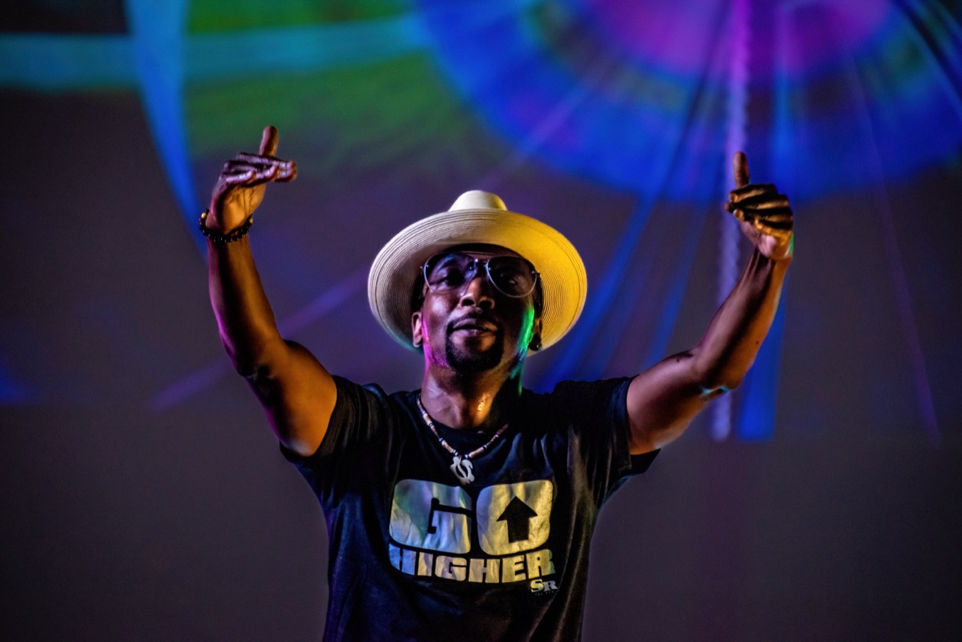 A performer in a wide straw hat lifts both hands, lit from below against a projection of sweeping blue, green and magenta light.