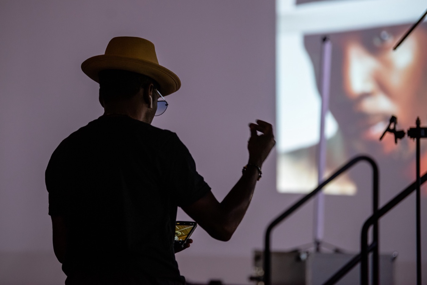 Shot from behind, a figure in a pale straw hat is outlined dark against a lit wall, one hand raised with the fingers curled. A large projected image of a face fills the wall to the right.
