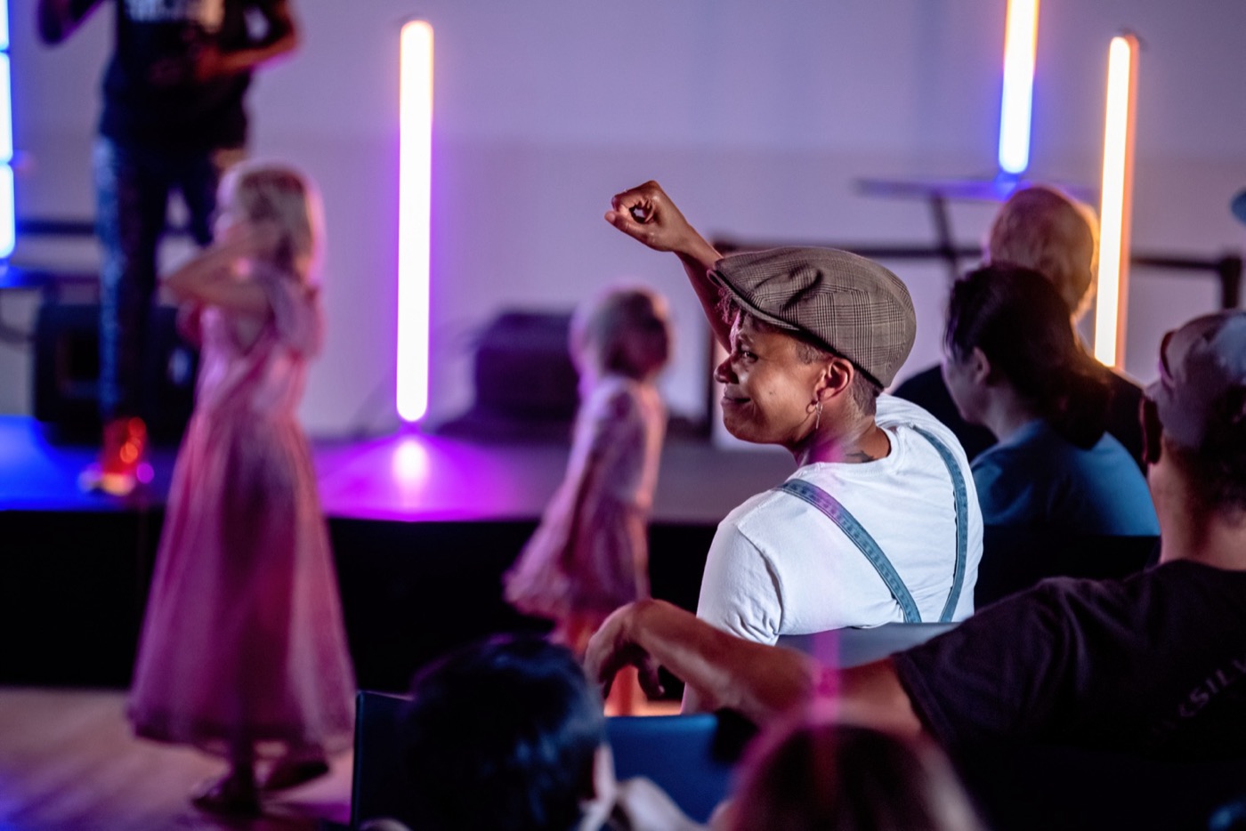 An audience member in a flat cap raises a fist from a seated row, looking toward the stage. Orange and blue light tubes stand behind, and a figure in a long dress is caught out of focus at the edge of the frame.