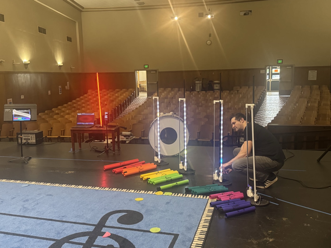 A school auditorium stage set for a workshop: tall LED tubes lit in color, a bass drum, and a row of color coded percussion tubes laid out across a floor mat printed with a musical staff, with rows of empty seats behind.