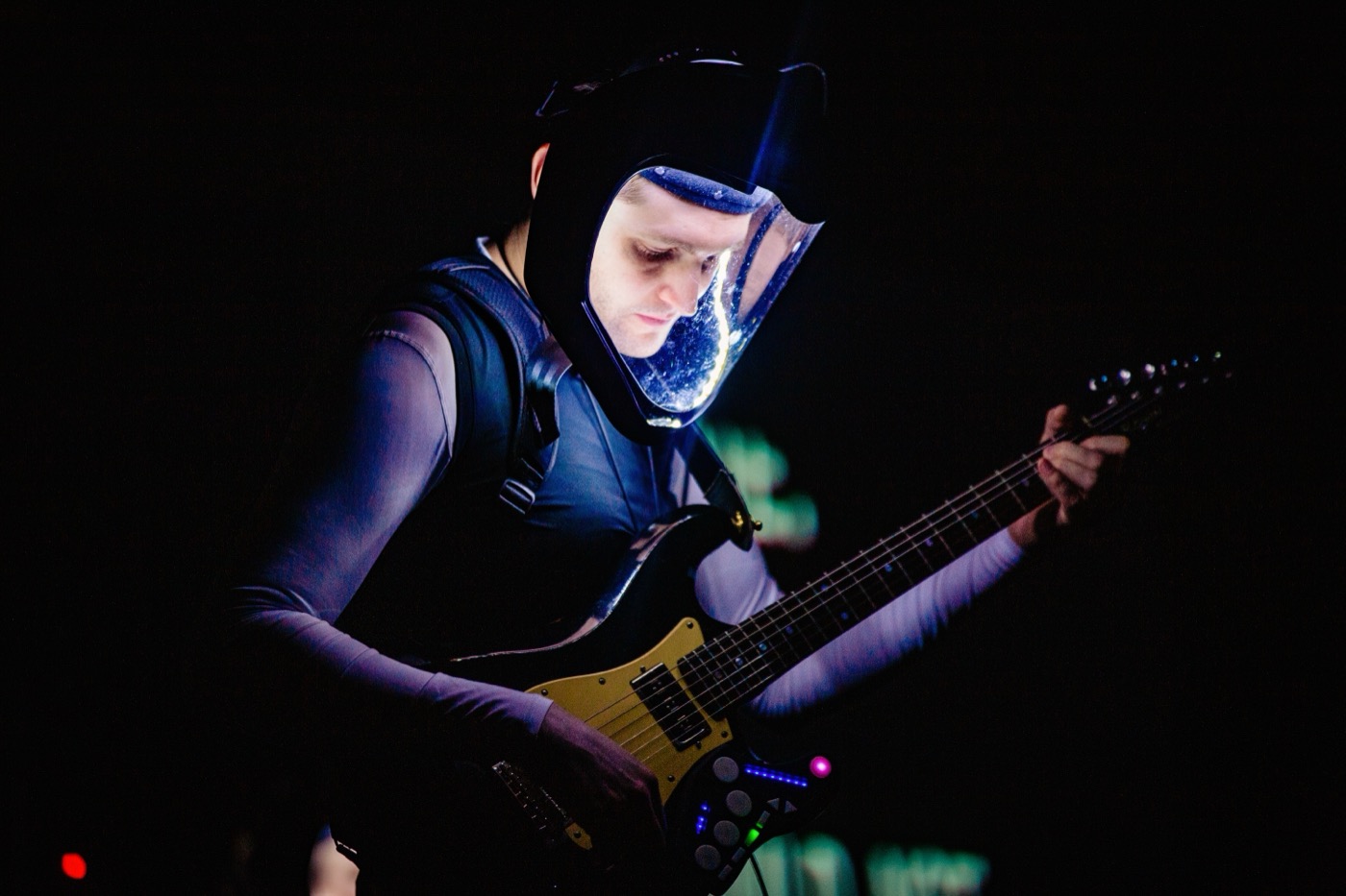 Close view of a guitarist in a lit-up helmet under deep blue and violet light, wearing a vest covered in small devices, with extra controls fixed to the guitar.
