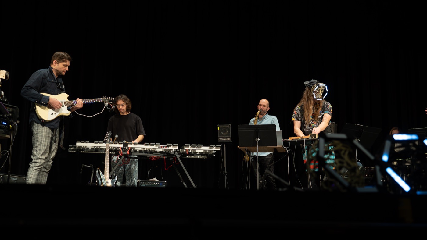 Four players spread across a dark stage with a guitar, a mallet instrument, a saxophone and a table of electronics, lit wearable devices glowing near the front of the frame.
