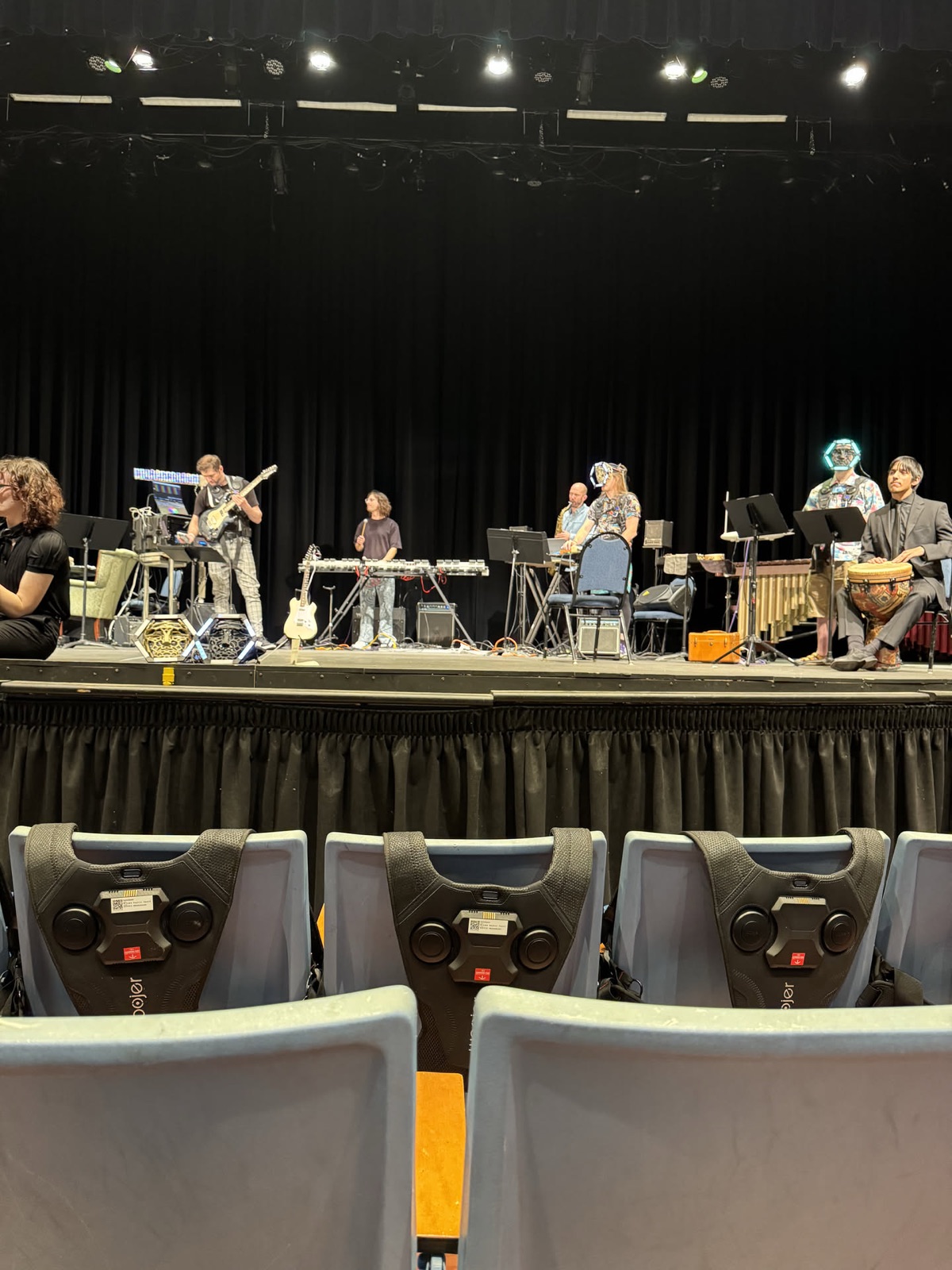 An ensemble at instruments across a lit stage, seen from the back of the hall over rows of empty auditorium seats.