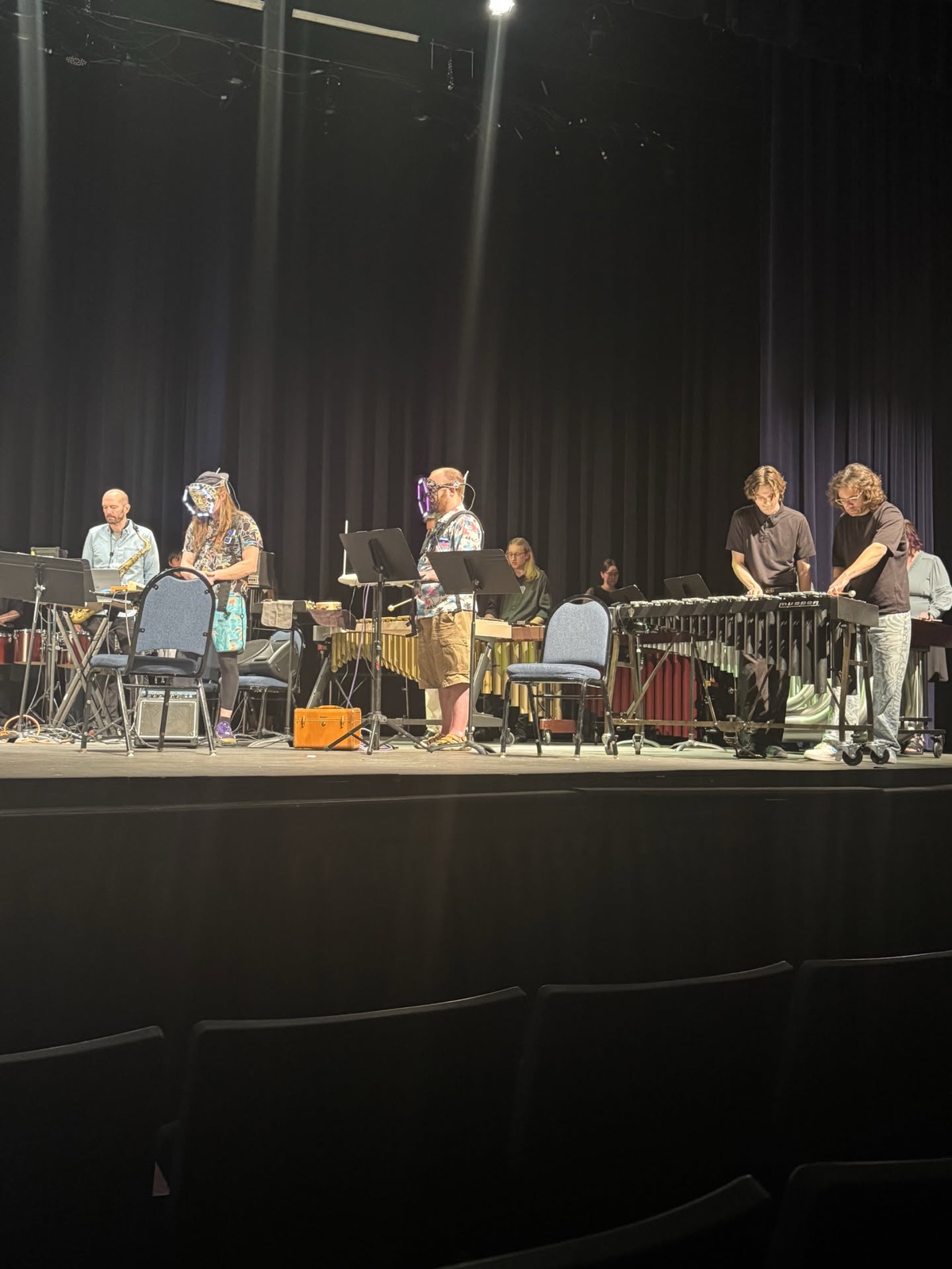 About ten people spread across a stage playing marimbas, vibraphone, hand drums, keyboards and guitars together, several wearing lit headgear and haptic vests.