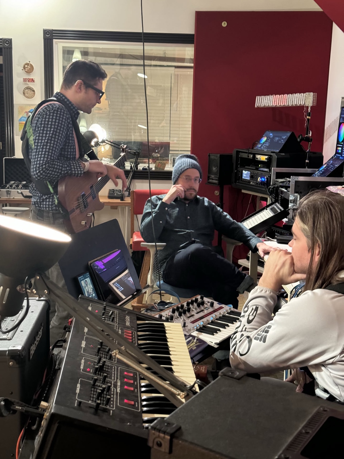 Three people working closely at a bank of synthesizers and screens, one playing a bass, one seated listening, one leaning in at the keys.