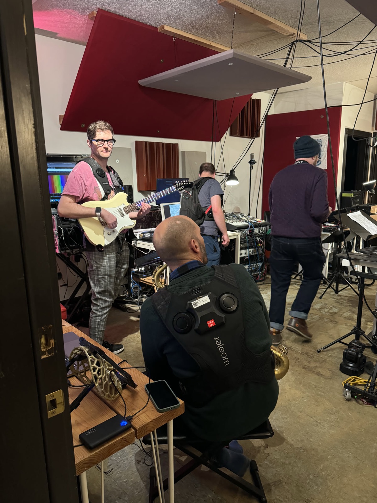 A working session in a rehearsal room: a guitarist plays and laughs while a person in the foreground wears a haptic vest with round drivers across the back, and two others set up gear behind them.
