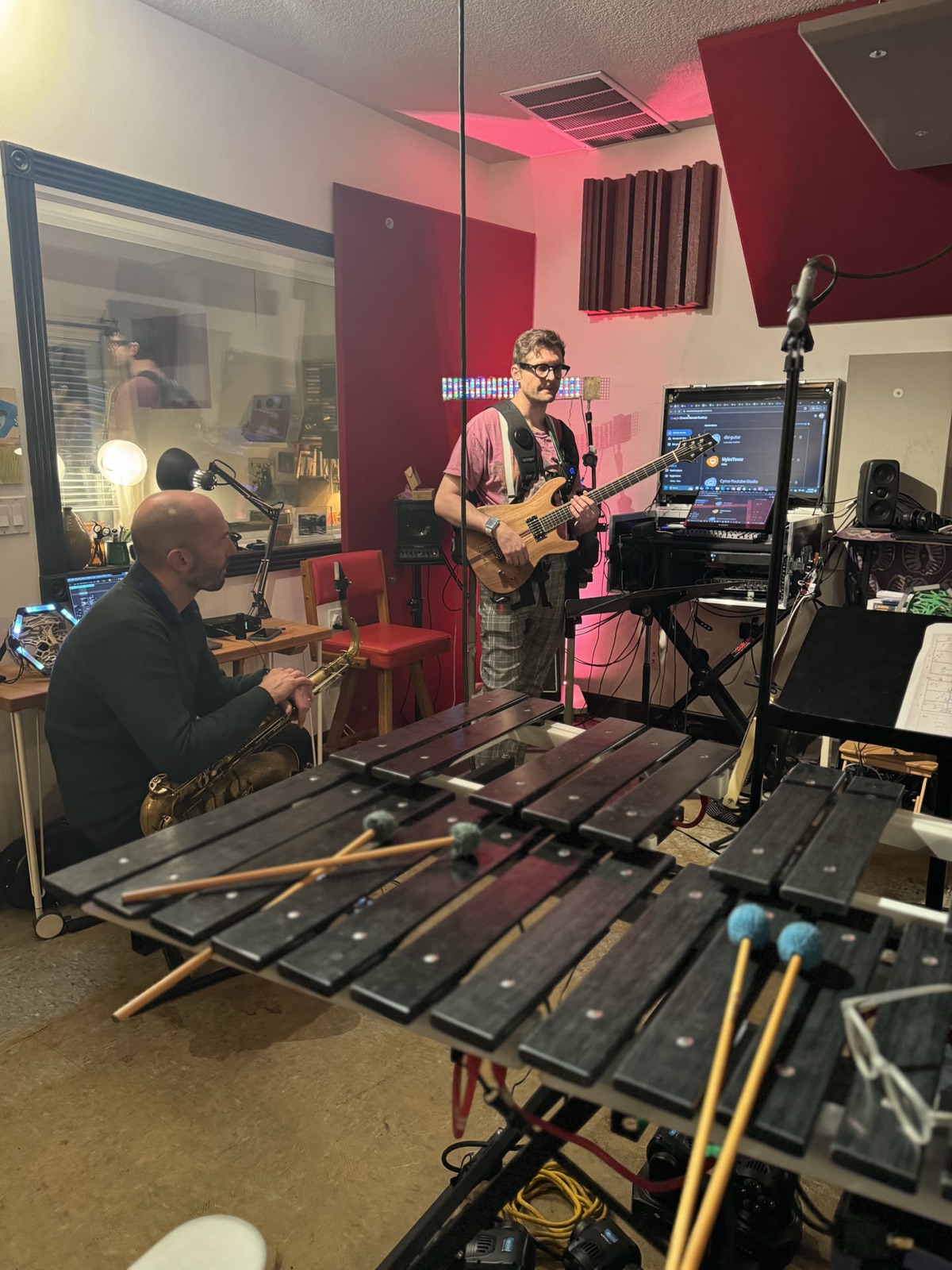 Two musicians at a vibraphone, an older man seated with a saxophone across his lap reaching in with a mallet while a standing guitarist plays beside him.