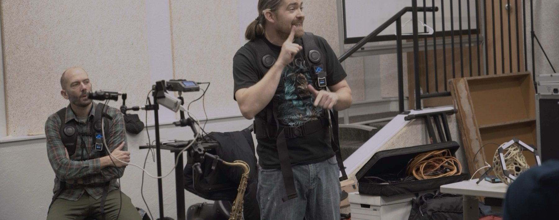 A workshop leader signs mid-session, wearing a haptic vest, with a saxophone, a camera and recording equipment around him in a university rehearsal room.